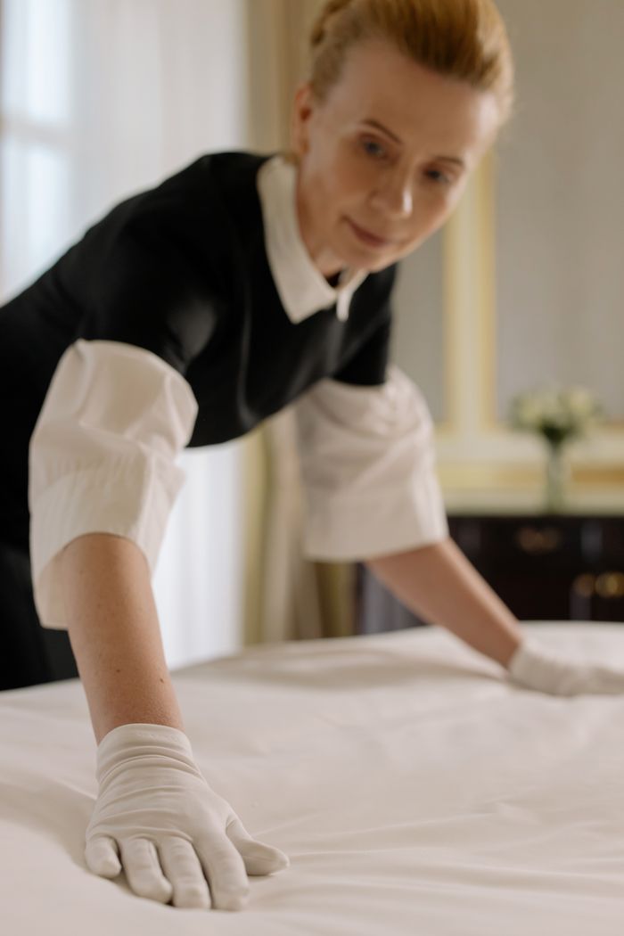 who-we-are Une femme de chambre dispose avec élégance les draps dans une chambre d’hôtel luxueuse, mettant en valeur son expertise en hôtellerie.